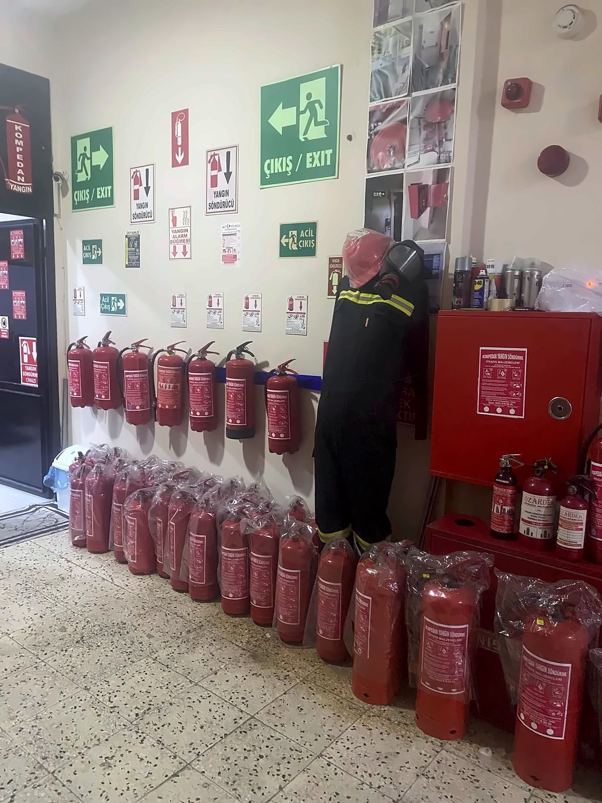 Kompedan dükkânının içi: duvarda asılı yangın söndürme tüpleri, acil çıkış ve yangın söndürücü levhaları, zeminde poşet içinde sıralanmış tüpler, itfaiye kıyafeti giydirilmiş manken ve kırmızı yangın dolabı